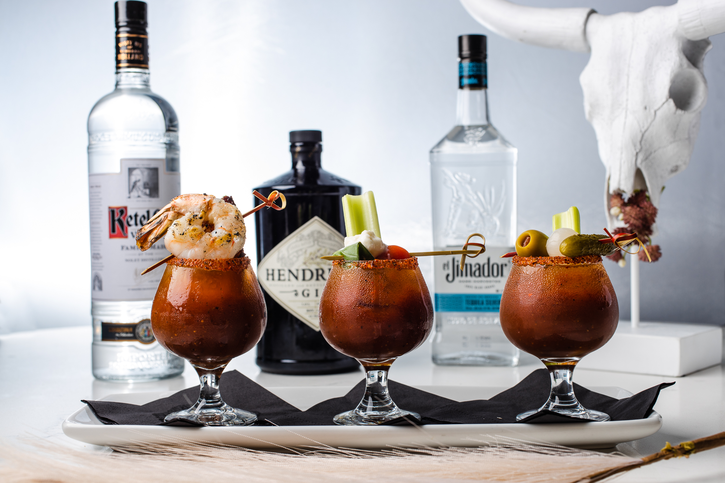 Three garnished Bloody Mary cocktails with liquor bottles behind them