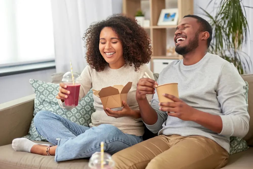 A couple laughing together on their couch while eating restaurant takeout at home