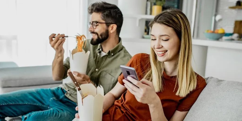 A couple relaxing at home with takeout containers while one of them places an order on a smartphone
